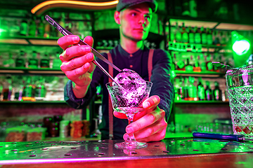 Image showing Close up of barman finishes preparation of alcoholic cocktail, pouring drink in multicolored neon light, focus on glass