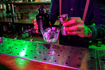 Image showing Close up of barman finishes preparation of alcoholic cocktail, pouring drink with shot in multicolored neon light, focus on glass