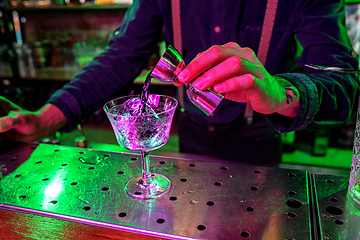 Image showing Close up of barman finishes preparation of alcoholic cocktail, pouring drink with shot in multicolored neon light, focus on glass