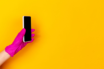 Image showing Hand in pink rubber glove holding smartphone with blank screen isolated on yellow studio background with copyspace.