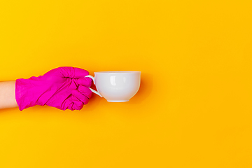 Image showing Hand in pink rubber glove holding coffee cup isolated on yellow studio background with copyspace.