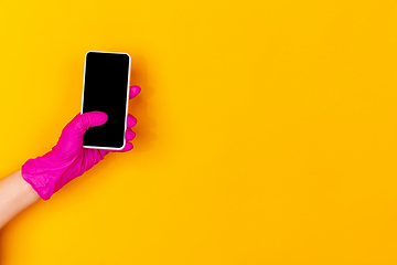 Image showing Hand in pink rubber glove holding smartphone with blank screen isolated on yellow studio background with copyspace.