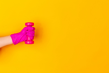 Image showing Hand in pink rubber glove holding dumbbell, gym weight isolated on yellow studio background with copyspace.