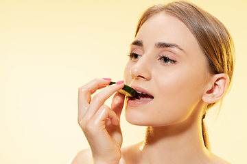 Image showing Biting. Close up of beautiful female face with cucumber slice over yellow background. Cosmetics and makeup, natural and eco treatment, skin care.
