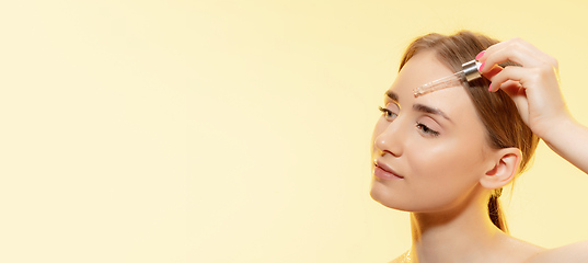 Image showing Clear. Close up of beautiful female face with pouring essential oil, serum over yellow background. Cosmetics and makeup, natural and eco treatment, skin care. Flyer with copyspace.