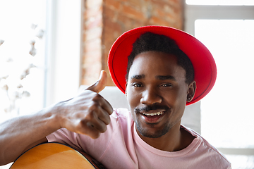 Image showing African-american musician in red hat greeting audience before online concert at home isolated and quarantined, cheerful, smiling