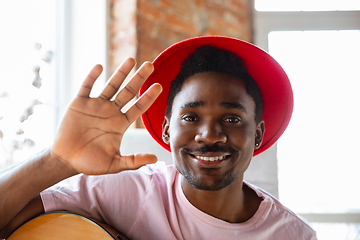 Image showing African-american musician in red hat greeting audience before online concert at home isolated and quarantined, cheerful, smiling