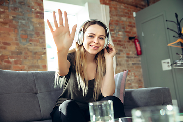 Image showing Young woman wearing wireless headphones gesturing during a video conference in the living room. Waving