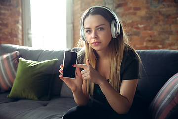 Image showing Young woman wearing wireless headphones gesturing during a video conference in the living room. Pointing on phone