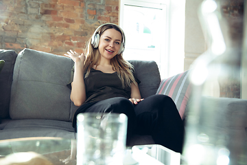 Image showing Young woman wearing wireless headphones gesturing during a video conference in the living room. Waving