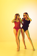 Image showing Beautiful girls in fashionable swimsuits isolated on yellow studio background in neon light. Summer, resort, fashion and weekend concept.