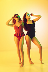 Image showing Beautiful girls in fashionable swimsuits isolated on yellow studio background in neon light. Summer, resort, fashion and weekend concept.