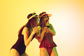 Image showing Beautiful girls in fashionable swimsuits isolated on yellow studio background in neon light. Summer, resort, fashion and weekend concept. Drinking cocktails.