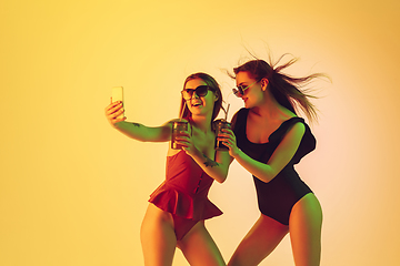 Image showing Beautiful girls in fashionable swimsuits isolated on yellow studio background in neon light. Summer, resort, fashion and weekend concept. Taking selfie with cocktails.