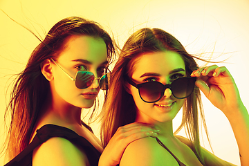 Image showing Close up beautiful girls in fashionable swimsuits and eyewear isolated on yellow studio background in neon light. Summer, resort, fashion and weekend concept.