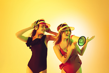 Image showing Beautiful girls in fashionable swimsuits and eyewear, headphones isolated on yellow studio background in neon light. Summer, resort, fashion and weekend concept. Calling with megaphone.