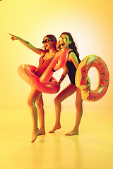 Image showing Beautiful girls in fashionable swimsuits isolated on yellow studio background in neon light. Summer, resort, fashion and weekend concept. Posing in rubber donut and flamingo with bright emotions.