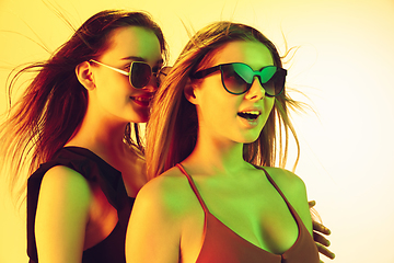 Image showing Close up beautiful girls in fashionable swimsuits and eyewear isolated on yellow studio background in neon light. Summer, resort, fashion and weekend concept.