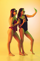 Image showing Beautiful girls in fashionable swimsuits isolated on yellow studio background in neon light. Summer, resort, fashion and weekend concept. Taking selfie with cocktails.