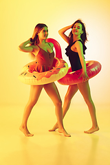 Image showing Beautiful girls in fashionable swimsuits isolated on yellow studio background in neon light. Summer, resort, fashion and weekend concept. Posing in rubber donut and flamingo with bright emotions.