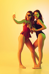 Image showing Beautiful girls in fashionable swimsuits isolated on yellow studio background in neon light. Summer, resort, fashion and weekend concept. Taking selfie with cocktails.