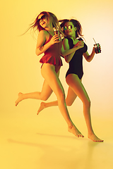 Image showing Beautiful girls in fashionable swimsuits isolated on yellow studio background in neon light. Summer, resort, fashion and weekend concept. With cocktails on the run.