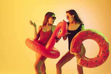 Image showing Beautiful girls in fashionable swimsuits isolated on yellow studio background in neon light. Summer, resort, fashion and weekend concept. Posing in rubber donut and flamingo with bright emotions.