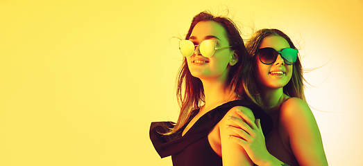 Image showing Close up beautiful girls in fashionable swimsuits and eyewear isolated on yellow studio background in neon light. Summer, resort, fashion and weekend concept. Flyer with copyspace.