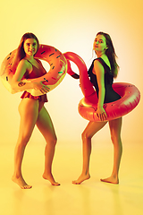 Image showing Beautiful girls in fashionable swimsuits isolated on yellow studio background in neon light. Summer, resort, fashion and weekend concept. Posing in rubber donut and flamingo with bright emotions.