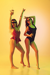 Image showing Beautiful girls in fashionable swimsuits isolated on yellow studio background in neon light. Summer, resort, fashion and weekend concept. Dancing with cocktails.