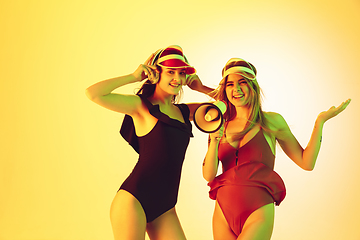 Image showing Beautiful girls in fashionable swimsuits and eyewear, headphones isolated on yellow studio background in neon light. Summer, resort, fashion and weekend concept. Calling with megaphone.