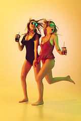 Image showing Beautiful girls in fashionable swimsuits isolated on yellow studio background in neon light. Summer, resort, fashion and weekend concept. With cocktails on the run.