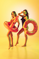 Image showing Beautiful girls in fashionable swimsuits isolated on yellow studio background in neon light. Summer, resort, fashion and weekend concept. Posing in rubber donut and flamingo with bright emotions.