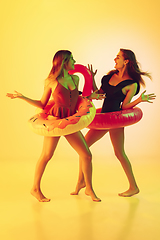 Image showing Beautiful girls in fashionable swimsuits isolated on yellow studio background in neon light. Summer, resort, fashion and weekend concept. Posing in rubber donut and flamingo with bright emotions.