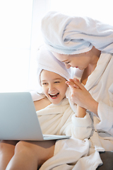 Image showing Mother and daughter, sisters have quite, beauty and fun day together at home. Comfort and togetherness. Watching series using laptop near window wearing white bathrobes