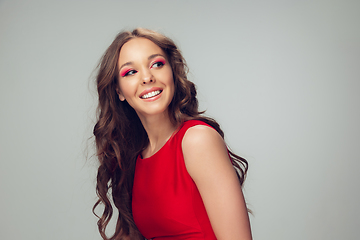 Image showing Beautiful young woman with long healthy curly hair and bright make up wearing red dress isolated on grey studio backgroud. Smiling.