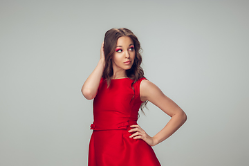 Image showing Beautiful young woman with long healthy curly hair and bright make up wearing red dress isolated on grey studio backgroud. Calm, thoughtful.