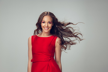 Image showing Beautiful young woman with long healthy curly hair and bright make up wearing red dress isolated on grey studio backgroud. Flying, blowing hair.