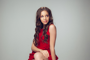 Image showing Beautiful young woman with long healthy curly hair and bright make up wearing red dress isolated on grey studio backgroud. Calm, thoughtful.