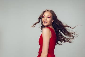 Image showing Beautiful young woman with long healthy curly hair and bright make up wearing red dress isolated on grey studio backgroud. Flying, blowing hair.