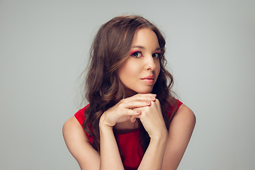 Image showing Beautiful young woman with long healthy curly hair and bright make up wearing red dress isolated on grey studio backgroud. Calm, thoughtful.