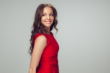Image showing Beautiful young woman with long healthy curly hair and bright make up wearing red dress isolated on grey studio backgroud. Smiling.