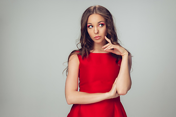 Image showing Beautiful young woman with long healthy curly hair and bright make up wearing red dress isolated on grey studio backgroud. Calm, thoughtful.