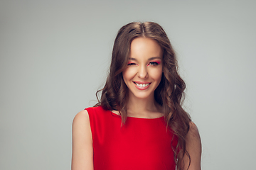 Image showing Beautiful young woman with long healthy curly hair and bright make up wearing red dress isolated on grey studio backgroud, winking, smiling