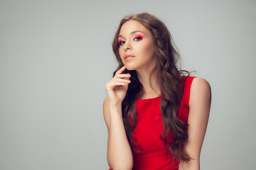 Image showing Beautiful young woman with long healthy curly hair and bright make up wearing red dress isolated on grey studio backgroud. Calm, thoughtful.