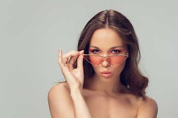 Image showing Close up of beautiful young woman with long healthy curly hair and bright make up wearing stylish pink eyewear isolated on grey studio backgroud, blowing