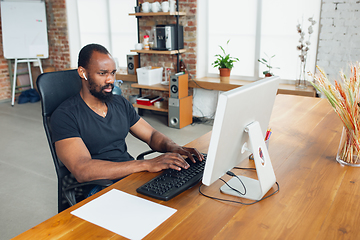 Image showing Young man, businessman working in office, looking on computer screen, monitor, with blank white sheet, whiteboard near him. Copyspace.