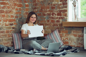 Image showing Young woman, businesswoman working or studying at home, looking on computer screen, monitor, holding white sheet, whiteboard. Attented, concentrated. Copyspace. Top view.