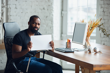 Image showing Young man, businessman working in office, looking on blank black computer screen, monitor, holding blank white sheet, whiteboard. Copyspace.