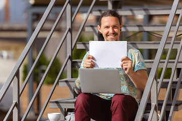 Image showing Young man, businessman working outdoors, looking on computer screen, monitor, holding blank white sheet, whiteboard. Copyspace.